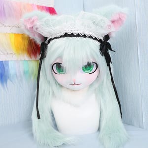 May include: A mint green furry cosplay head with large green eyes, a white face, and pink cheeks. The head is adorned with a white lace bonnet with black trim and bows. Long mint green fur extends from the head.
