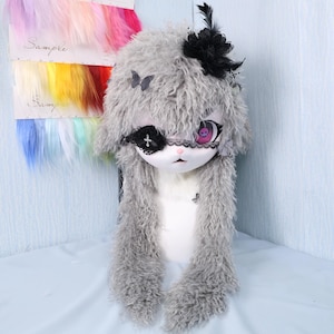 May include: A plush, gray bunny doll with long, shaggy fur, adorned with a black eye patch, a skull-themed eye, and a black flower. The doll has a white face and chest, with butterfly and cross accents. Colorful hair samples are visible in the background.