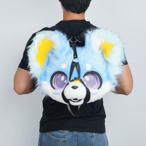 May include: A plush backpack shaped like a stylized animal head with long light blue faux fur, bright yellow inner ears, and large purple eyes. The bag features a white muzzle, a black nose, and black straps that secure the head-shaped compartment.