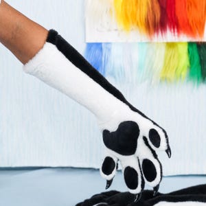 May include: A long, faux fur glove designed as an animal paw, featuring a black and white color scheme. The glove includes a large black paw pad, four smaller black toe pads, and sharp black claws extending from each of the four fingers.