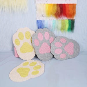 May include: Four paw-shaped mats in cream and gray, with yellow and pink paw pads. The mats have a textured surface. A swatch of colorful faux fur and a sample card are in the background.