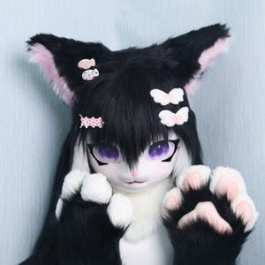 May include: A black and white cat fursuit head with large purple eyes and pink accents. The ears have pink inner fur, and the paws are black with pink pads. Small decorative hair clips adorn the head.