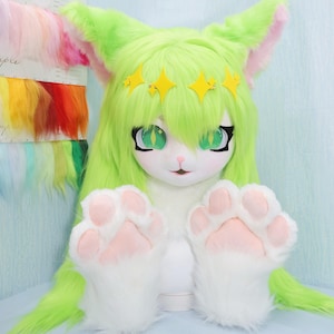 May include: A cat-like costume head in white and green, featuring large green eyes and a yellow star crown. The costume has long, green hair and large, fluffy white paws with pink pads. Designed for wear, it's a striking cosplay piece.