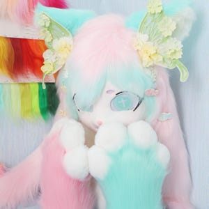Peut inclure: Un costume de fourrure aux couleurs pastel, comprenant une tête et des pattes. La tête présente un visage blanc avec de grands yeux, des cheveux bleus et roses, et des ornements floraux. Les pattes sont un mélange de fourrure blanche et bleue.