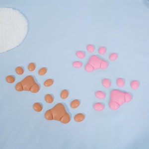May include: Two sets of paw prints, one in light brown and the other in pink, are arranged on a light blue background. Each paw print has three larger pads and four smaller toe pads. A white, fluffy, oval shape is in the upper left corner.