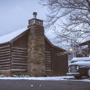 May include: A black vintage pickup truck covered in snow is parked in front of a rustic log cabin with a stone chimney. The cabin has a snow-covered roof and is surrounded by trees.