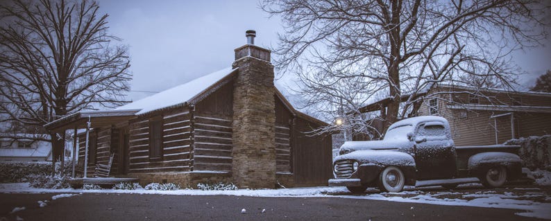 May include: A black vintage pickup truck covered in snow is parked in front of a rustic log cabin with a stone chimney. The cabin has a snow-covered roof and is surrounded by trees.
