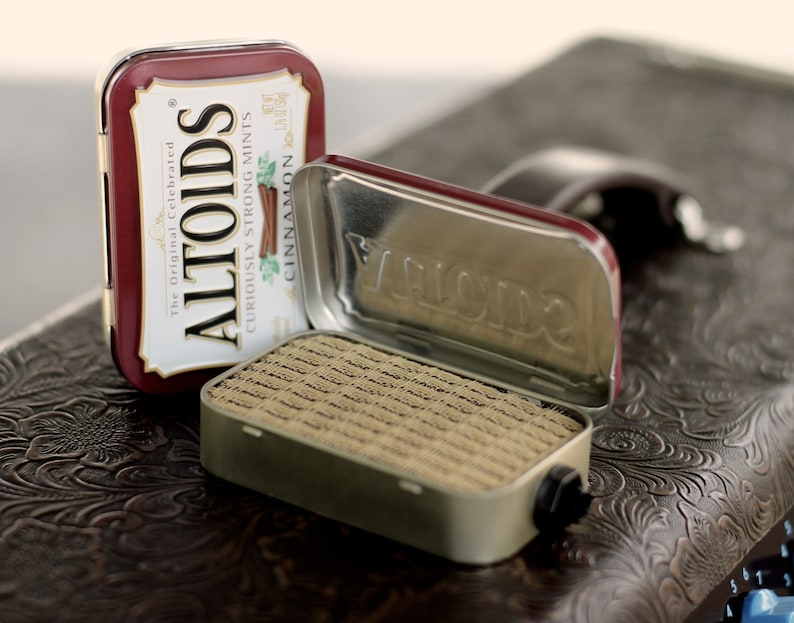 May include: Two Altoids tins, one open and one closed. The closed tin is red with white lettering that reads "Altoids, The Original Celebrated Curiously Strong Mints, Cinnamon". The open tin is silver with a brown, rectangular mint container inside.
