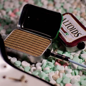 May include: A small, silver, rectangular speaker with a brown speaker grille sits on a bed of white and green candy mints. A silver cable with a black plug is connected to the speaker. A red and white Altoids mint tin is also visible in the image.