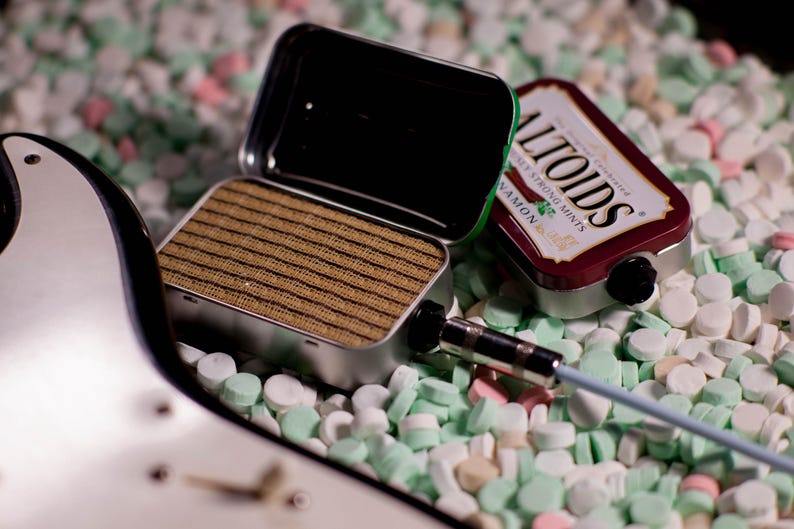 May include: A small, silver, rectangular speaker with a brown speaker grille sits on a bed of white and green candy mints. A silver cable with a black plug is connected to the speaker. A red and white Altoids mint tin is also visible in the image.