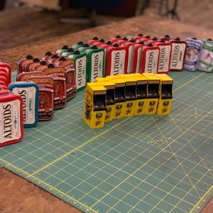 May include: A collection of miniature arcade cabinets and Altoids tins. The arcade cabinets are yellow with black details and the tins are red, green, and blue with the word "Altoids" printed on them.