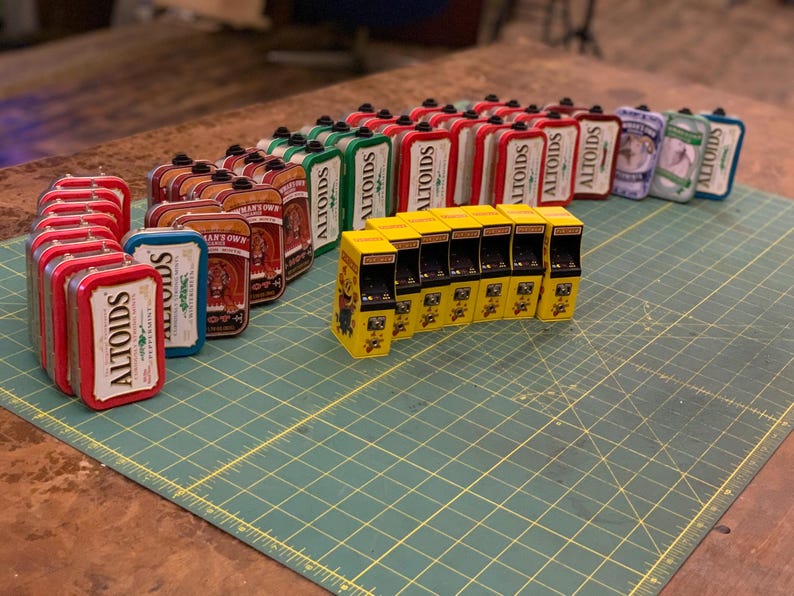 May include: A collection of miniature arcade cabinets and Altoids tins. The arcade cabinets are yellow with black details and the tins are red, green, and blue with the word "Altoids" printed on them.