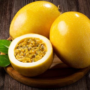 Puede incluir: Tres maracuyás amarillas, una cortada por la mitad para revelar la pulpa, descansan sobre un plato de madera con hojas verdes. Las frutas tienen una piel lisa y brillante. El fondo es una superficie de madera oscura.
