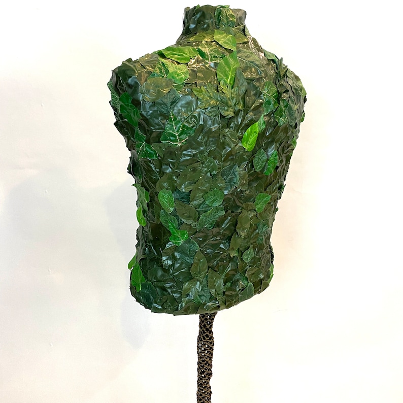 May include: A torso-shaped mannequin covered in green leaves. The leaves are a mix of light and dark green, creating a textured surface. The mannequin is mounted on a brown, textured stand, and set against a white background.