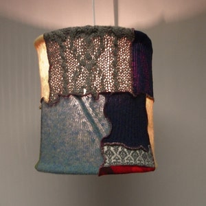 May include: A patchwork lampshade made from different types of knitted fabric in shades of gray, blue, and red.
