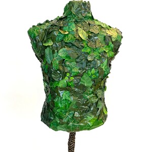 May include: A green mannequin torso covered in artificial leaves. The leaves vary in size and shade, creating a textured, natural appearance. The mannequin is mounted on a brown and gold patterned stand, set against a white background.