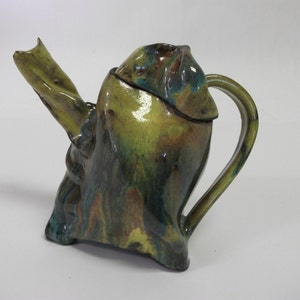 May include: A ceramic teapot with a unique, abstract design. The teapot is glazed in shades of green, blue, and brown, with a textured surface. The spout is twisted and the handle is curved.