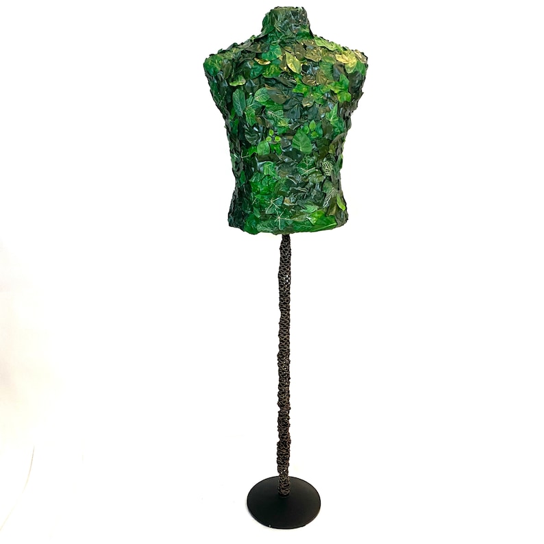 May include: A decorative mannequin torso covered in green leaves, mounted on a dark metal stand. The stand has a round base and a textured, twisted stem. The leaves vary in shades of green, creating a natural, organic aesthetic.