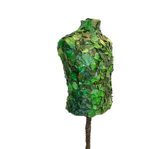 May include: A torso-shaped mannequin covered in green artificial leaves. The leaves vary in shade and size, creating a textured, natural appearance. The mannequin is mounted on a brown, textured stand, suitable for display or decorative purposes.