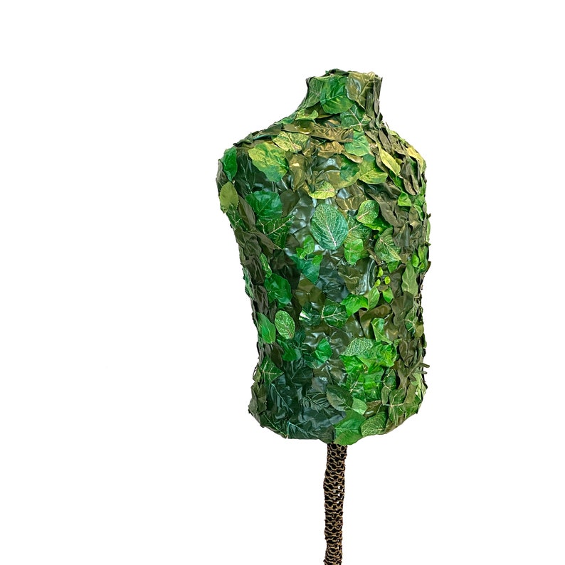 May include: A torso-shaped mannequin covered in green artificial leaves. The leaves vary in shade and size, creating a textured, natural appearance. The mannequin is mounted on a brown, textured stand, suitable for display or decorative purposes.
