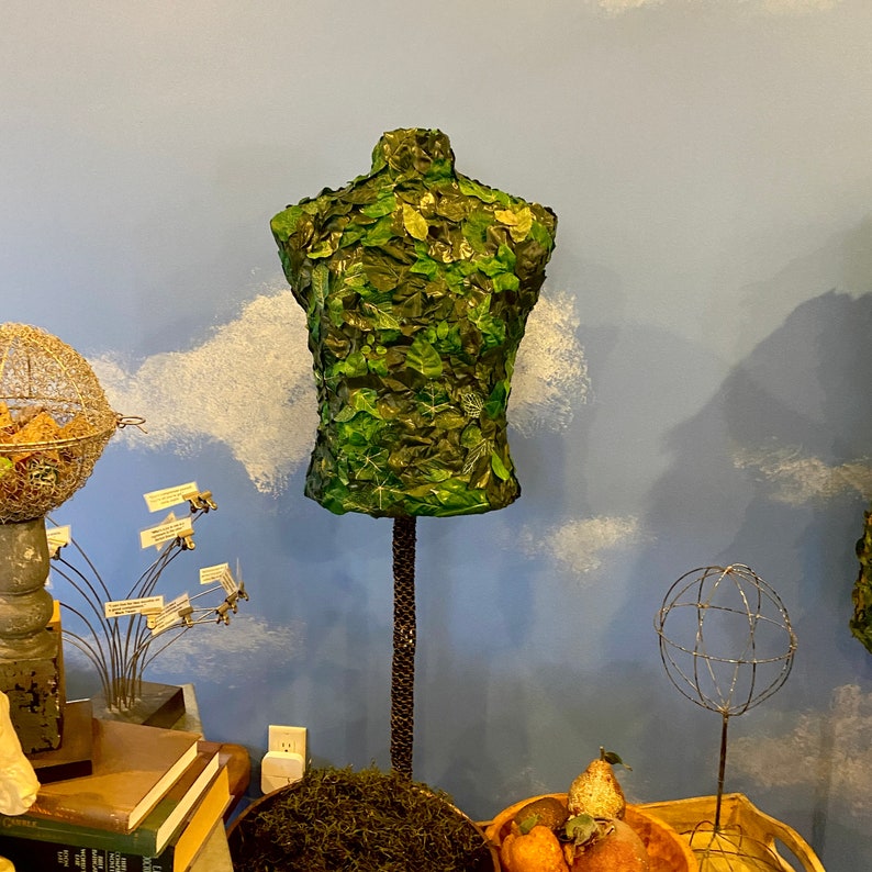 May include: A decorative torso form covered in green leaves, standing on a dark brown metal stand. The form is a vibrant green with detailed leaf patterns. The stand is supported by a round base of moss. The background is a light blue wall with white cloud designs.