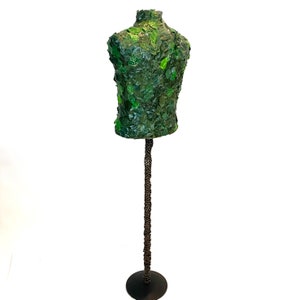 May include: A decorative torso mannequin covered in green leaves. The upper body form is mounted on a dark metal stand with a circular base. The leaves are various shades of green.
