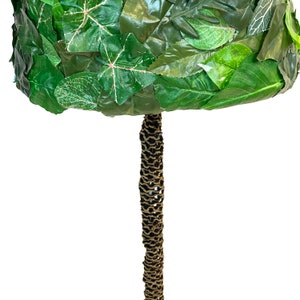 May include: A decorative topiary tree with a round, leafy crown made of green leaves. The trunk is wrapped in a textured, brown material. The leaves have a variety of green shades and some have pink veins.