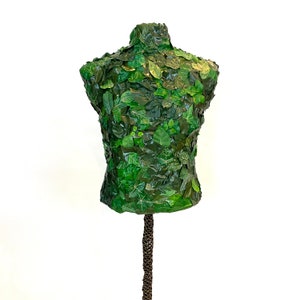 May include: A green mannequin torso covered in various sizes and shades of green leaves. The leaves create a textured surface, giving the mannequin a natural, organic appearance. The mannequin is mounted on a brown, textured stand, set against a white background.
