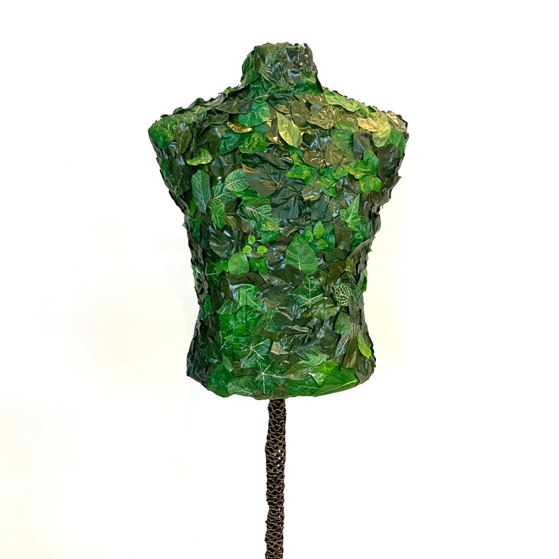 May include: A green mannequin torso covered in various sizes and shades of green leaves. The leaves create a textured surface, giving the mannequin a natural, organic appearance. The mannequin is mounted on a brown, textured stand, set against a white background.