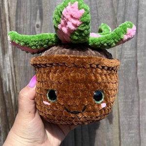May include: A handmade, smiling crochet succulent in a brown pot. The succulent has green and pink leaves. The pot has a smiling face with black eyes and pink cheeks. The item is held by a hand.