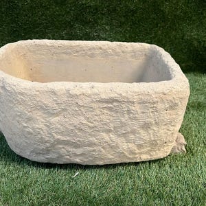 May include: A rectangular, beige planter with a textured, stone-like appearance. The planter has a wide, open top and sits on a green surface. The planter has small decorative feet.
