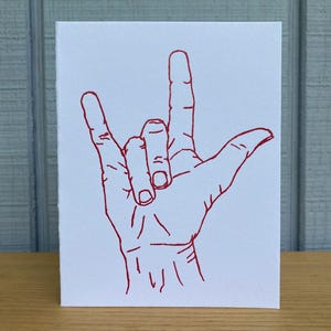 May include: A white greeting card with a red line drawing of a hand making the "I love you" sign in American Sign Language. The card is on a wooden surface.