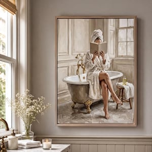 May include: A framed wall art print depicting a person in a white bathrobe and towel turban sitting on the edge of a clawfoot bathtub, holding an open book. The scene features a gold faucet, a small wooden stool, and a neutral-toned bathroom.