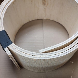 May include: A rolled-up, light-colored wooden veneer, held together with black tape. The wood grain is visible, and the layers are clearly defined. The shape suggests it could be used for crafting or construction projects.
