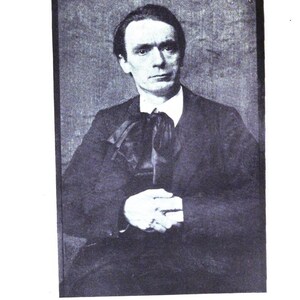 May include: A black and white portrait of a man in a dark suit with a white collared shirt and a large bow tie. The man is looking directly at the camera with his hands clasped in front of him. The background is a solid, neutral color.