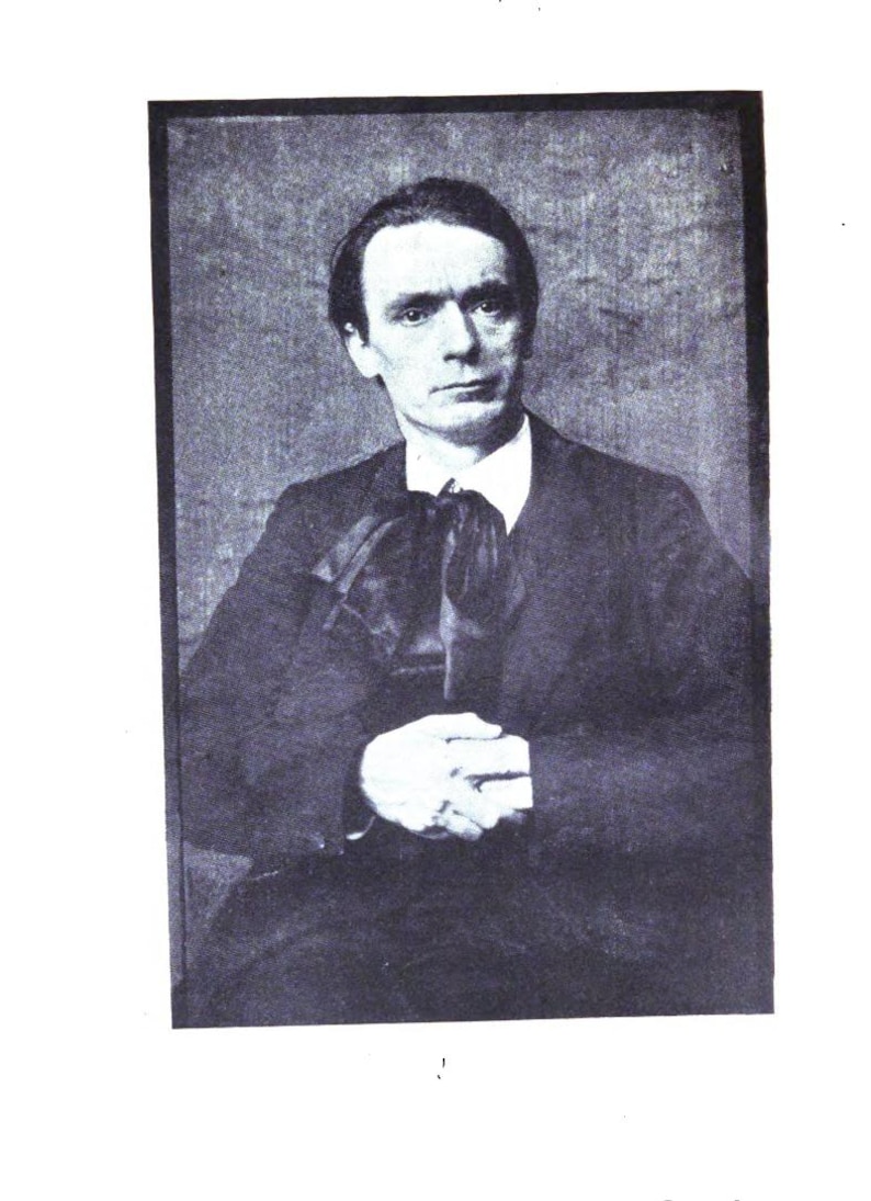 May include: A black and white portrait of a man in a dark suit with a white collared shirt and a large bow tie. The man is looking directly at the camera with his hands clasped in front of him. The background is a solid, neutral color.