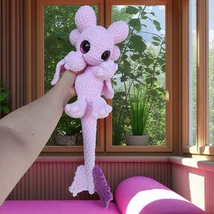 May include: A handmade, light pink crochet plush toy with large, dark pink eyes and a long, textured tail. The toy has a whimsical design with rounded features and small, petal-like appendages. The background includes a window with a view of greenery.