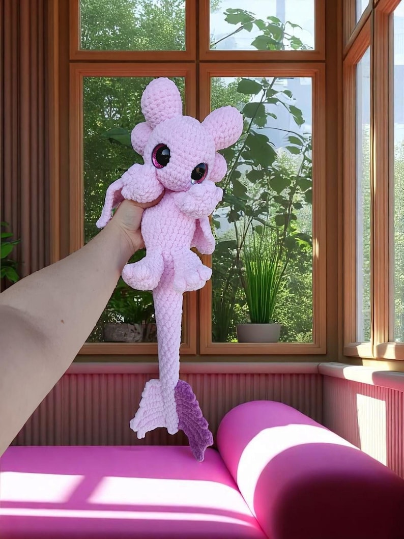 May include: A handmade, light pink crochet plush toy with large, dark pink eyes and a long, textured tail. The toy has a whimsical design with rounded features and small, petal-like appendages. The background includes a window with a view of greenery.