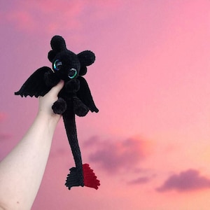 May include: A black crochet dragon plushie with large, iridescent eyes, wings, and a long tail with a red tip. The dragon is held up against a pink and purple sky background.