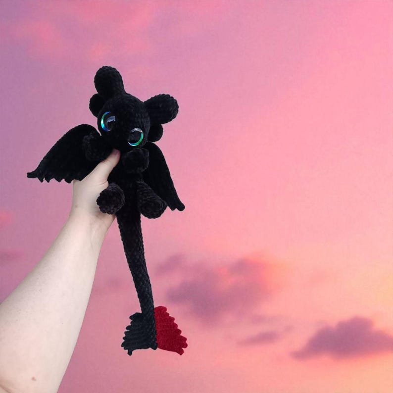 May include: A black crochet dragon plushie with large, iridescent eyes, wings, and a long tail with a red tip. The dragon is held up against a pink and purple sky background.