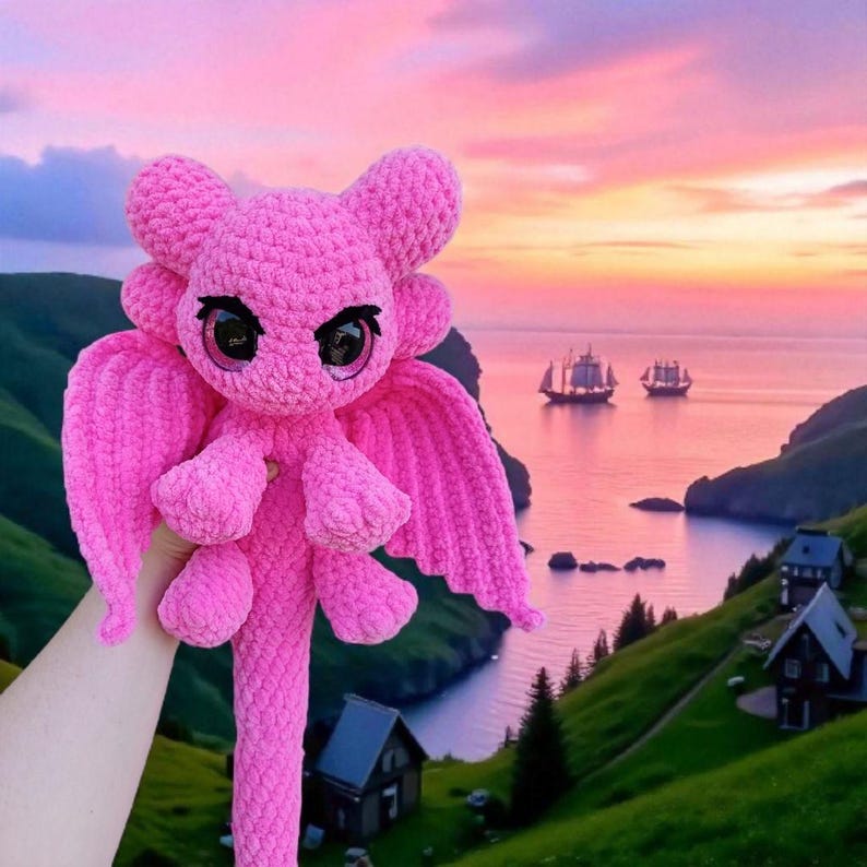 May include: A handmade, pink crochet dragon plushie with large, expressive eyes and wings. The dragon is held up against a scenic background of a sunset over the ocean with sailboats and rolling green hills.
