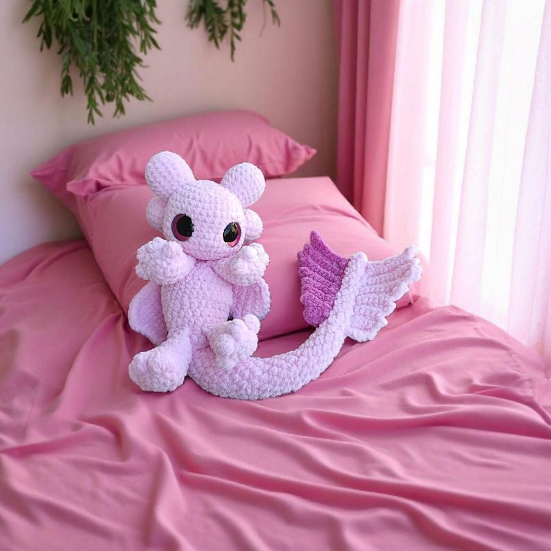 May include: A handmade, light pink crochet dragon plush toy with large, round eyes and a long tail. The dragon is sitting on a pink bed with a matching pillow and sheets. The tail has a purple and white fin. The background is a soft pink.