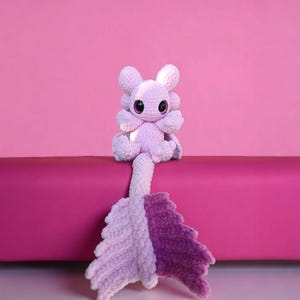 May include: A handmade, lavender-colored crochet plush toy in the shape of a mythical creature. It has large, black eyes, four arms, and a split tail in shades of lavender and purple. The toy is sitting on a magenta surface against a pink background.
