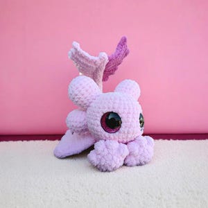 May include: A handmade, light purple crochet octopus plush toy with large, glittery black and pink eyes. The plush has four tentacles, two small ears, and two wing-like appendages in shades of pink and purple. The toy is on a white surface against a pink background.