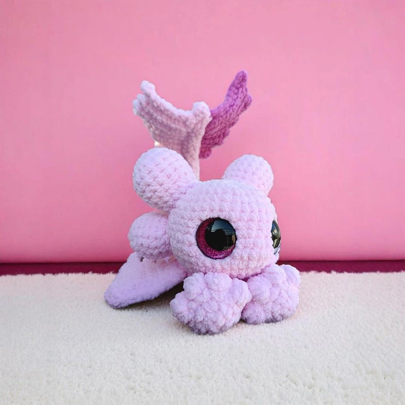 May include: A handmade, light purple crochet octopus plush toy with large, glittery black and pink eyes. The plush has four tentacles, two small ears, and two wing-like appendages in shades of pink and purple. The toy is on a white surface against a pink background.