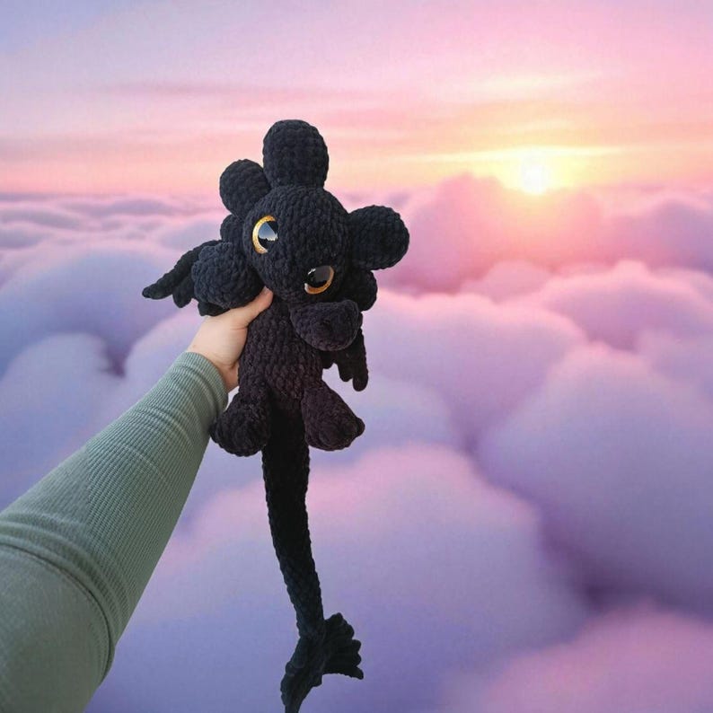 May include: A black crocheted dragon plush toy with large, golden eyes and small wings. The dragon has a long tail and is held up against a backdrop of a sunset over clouds. The plush is made of a soft, textured yarn.