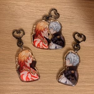 May include: Three acrylic keychains featuring anime-style illustrations of two characters. One character has red and orange hair, the other has white hair. Each keychain has a heart-shaped clasp.