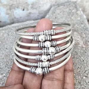 Può includere: Un gruppo di braccialetti d'argento. Ogni braccialetto presenta un design strutturato con dettagli di perline e una sfera centrale d'argento. I braccialetti sono esposti su uno sfondo neutro, evidenziando i loro dettagli.