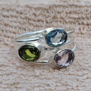 Può includere: Tre anelli d'argento, ciascuno con una pietra preziosa ovale. Uno ha una pietra azzurra, un altro una pietra verde e il terzo una pietra rosa-viola. Gli anelli sono esposti su una superficie strutturata bianco sporco.