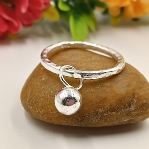 Puede incluir: Un anillo de plata con textura martillada y una esfera pulida colgante. El anillo se muestra sobre una piedra lisa y marrón, con flores borrosas en el fondo. El anillo es una joya.